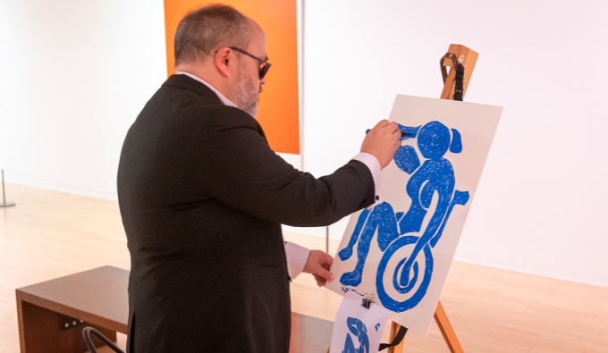 A man at an easel in a white gallery space wears a black suit and paints at an easel. The work is Matisse in style and depicts a person seated in a wheelchair. It is painted in blue against a white background. 