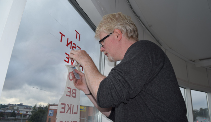 Aidan Moesby faces left toward a large window to which he is attaching red lettering. He wears dark rimmed glasses and a dark grey jumper