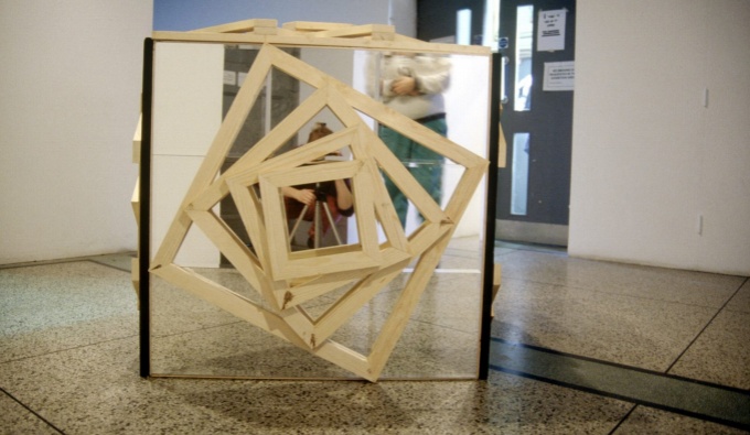 A mirrored square sculpture in a large room. Attached the face of the mirrored square facing the camera are several wooden square frames attached, every decreasing in size and leaving a small square at the centre of the mirror exposed.