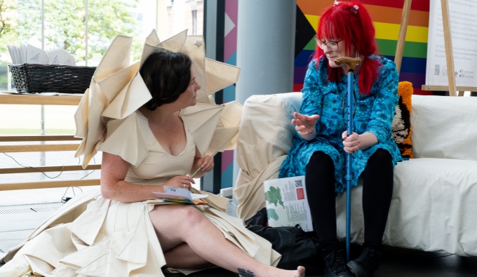 A woman with bright red hair and blue dress sits on a couch holding a walking stick. She addresses artist, Lisette who sits in front of her on the floor. Lisette where a cream dress of stiff fabric, with  pressed pleats around her shoulders and a train.