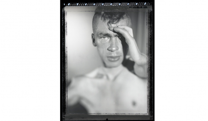 Black and white head and shoulders image of the great Mat Fraser. He is bare chested and is holding his left hand up to his forehead.  His head is shaved on both sides.