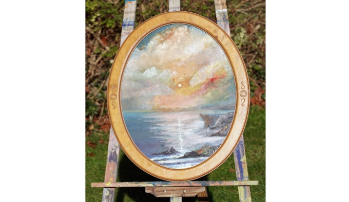 An oval cameo-style frame in gold containing a painting of a sunset over a bay, displayed on an easel.