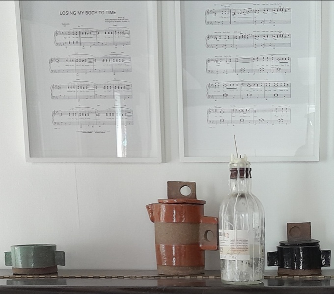 Two white framed pieces of music score, entitled: Losing my Body to Time. Below are two sculpted clay mugs with rectangular handles and a coffee pot in the same style, slightly off centre is a bottle with dried candle wax down the neck. 