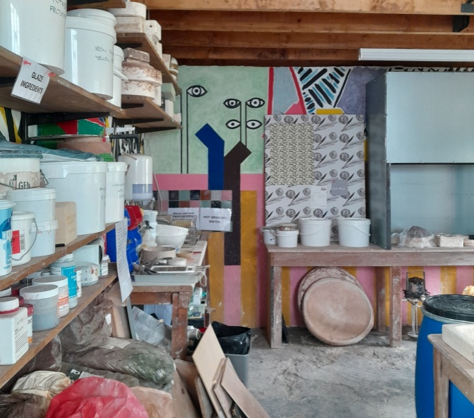 A photograph of a pottery studio. There are several shelves of glazes, clay etc to the left, and boards on the floor. The end wall has a bright abstract painting upon it. 