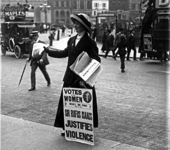 A British suffragette stans on the edge of the street handing out copies of the newspaper 'Votes for Women'. In front of her is a board with Votes for Women and 'Sir Rufus Isaacs Justifies Violence'. Behind her are trolley buses and people crossing the st