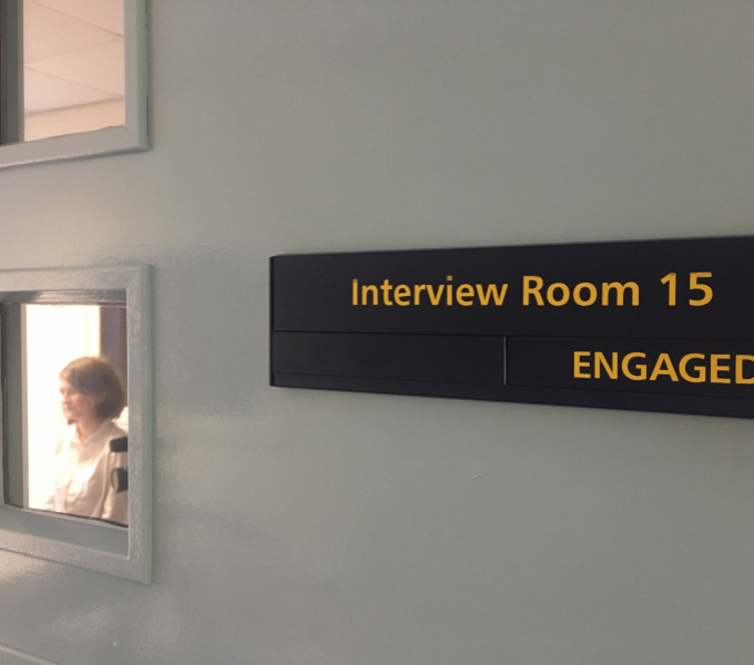 A photograph of a light grey painted wall with a large black rectangular sign on the right hand side of it with the words 'Interview Room 15, Engaged' on it in yellow. To the left are two small windows and through the lower window a woman is visible. 