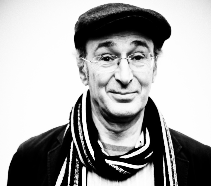 black and white photo of a clean shaven man, with a flat cap and striped scarf around his head.His dark eyebrows are raised as asking a question.
