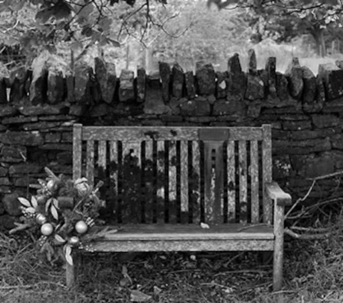 A black and white photographic image of an old wooden bench, in front of a dry stone wall. A bunch of now wilting flowers is tied to the arm of the bench.