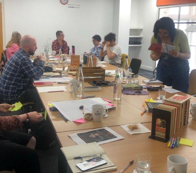 Six members of the DASh team are gathered around a large meeting table littered with books, pens, posters, water bottles and glasses and coffee cups. One person looks carefully at a book while others appear in comversation at the far end of the table. 