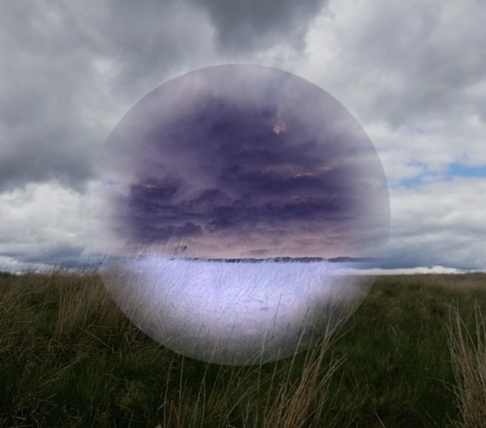 A landscape of green fields and purple cloudy sky. To the centre is a circular shape, partially obscuring the view, setting the background out of focus.