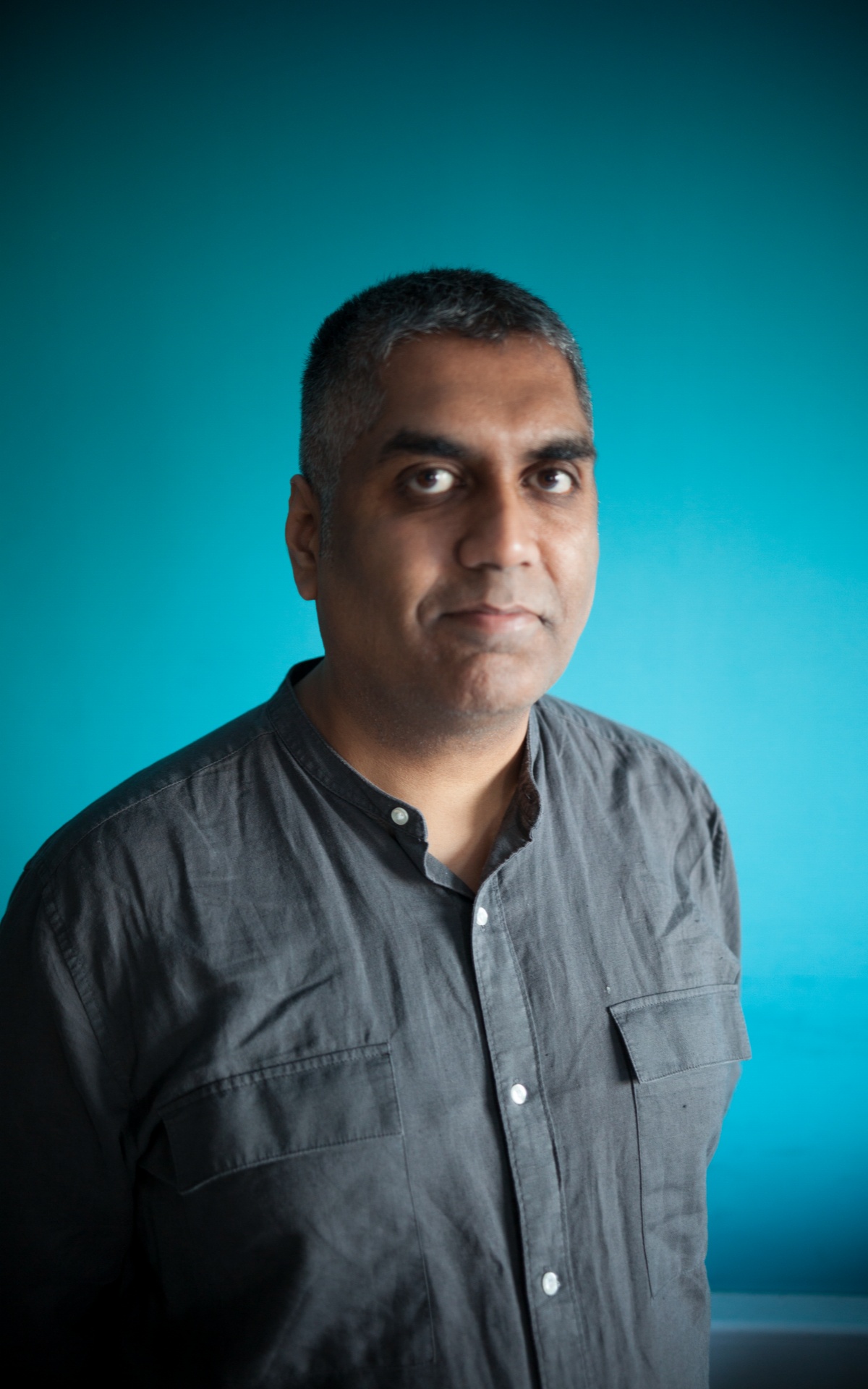 Ashok stands facing the camera against a turquoise background. He has short hair and wears a grey shirt. . He looks directly at the camera.