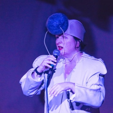 Nicola wears a light coloured jacket and a cap with sound equipment attached to the front of her head. It is shaped like a large pom pom. She holds the microphone to her lips as she addresses and audience. The room is lit with a purple, blue lighting. 