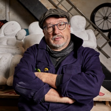 Tony Heaton in front of a large pale abstract sculpture. He wears a flat cap and glasses and dark blue overalls. His arms are folded as he looks at the camera.