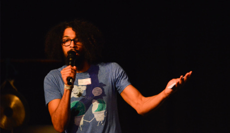 A black haired man in a blue and white t-shirt  hold the mike in his hand and raises his other arm and hand questioning 