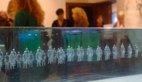 A photograph of a section of a long glass cuboid with many regularly spaced figures suspended upright within it. In the background, visitors observe the exhibition.