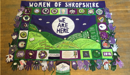 Banner created for the centenary of the 1st votes for women. 'WOMEN OF SHROPSHIRE' in felt letters at the top, 'WE ARE HERE' in felt letters, then the Shropshire hills in green felt below. Felt hands in purple, white and green surround the banner.
