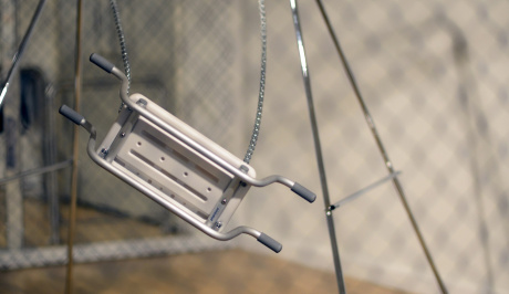 Through the blurred lines of close up netted fencing, a childs metal swing seems to be suspended in the air mid swing