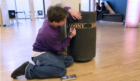 A young man is half sitting, half kneeling on the floor, dressed in blue jeans and and a purple jumper he is writing on a black,municipal type of litter bin, in gold capital letters  it spells out the word LOONY