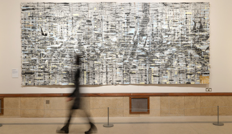 A black shadowy impression of a woman walks past a large wall, on the wall is a large painting in monochrome, white, greys and black, almost like a woven irregular pattern,as though marking changes in frequency.