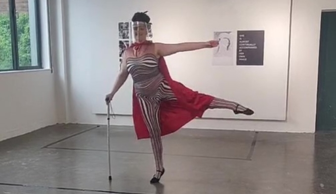 Porcelain dances in a white gallery space wearing a black and white striped leotard and a red cape. Their right arm holds a cane whilst the left arm and left leg are outstretched as she dances. 