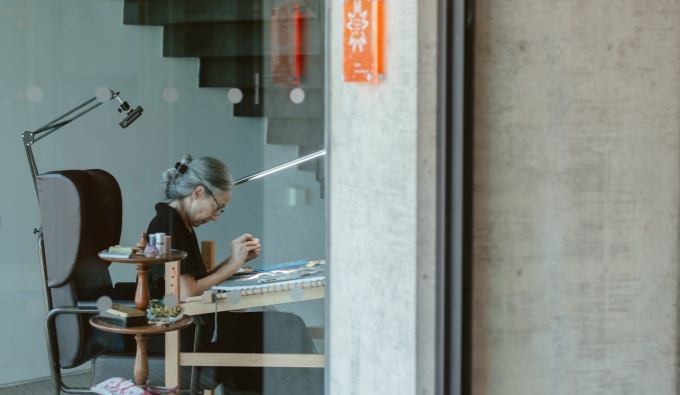 A view of Andrea through a large plate glass window. She is seated and works on her embroidery. 