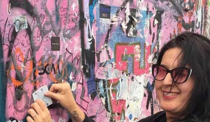 April has dark hair and wears dark rimmed glasses. She is to the right of the image and turns to face the camera as she smiles. She looks to be addinga stencil or a sticker to a large wall covered in graffiti and motifs