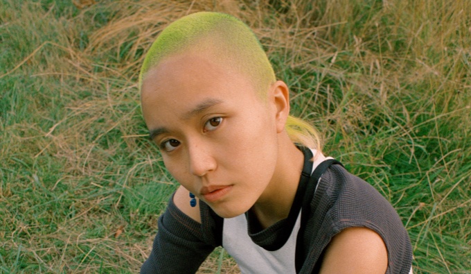 An image of April from the shoulders up. They are sat on grass and look towards the camera, they have shaved hair in a greenish colour and wear a black and white top.