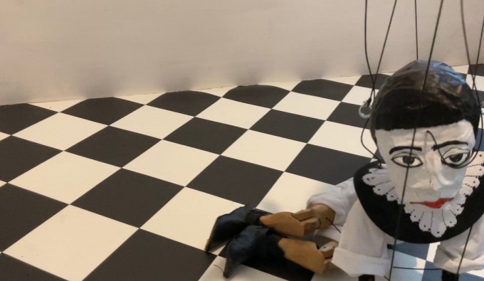 A black and white square tiled floor. To the right is a handmade puppet with a white face, black hat and and black and white costume. The puppet looks slightly upwards towards the camera.