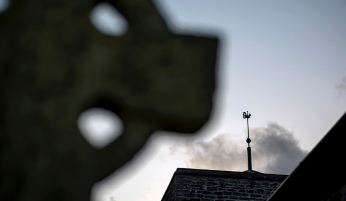 A view looking skywards, with the top of stone grave stone and partial rooftop of a church in silhouette.