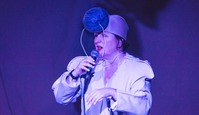 Nicola wears a white specially adapted 'Sound Suit' for her performance. On her head is a small tight hat with as large pom pom sound buffer. The stage is lit with blue lighting as she holds her microphone and performs to the audience.