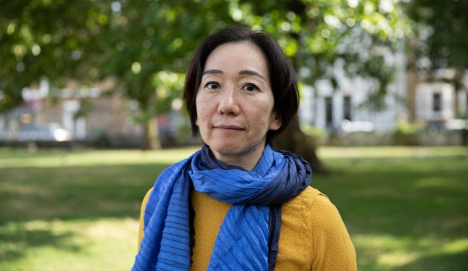 Chisato has short bobbed dark hair and wears a yellow top with a blue scarf. They stand in a park as the face the camera.