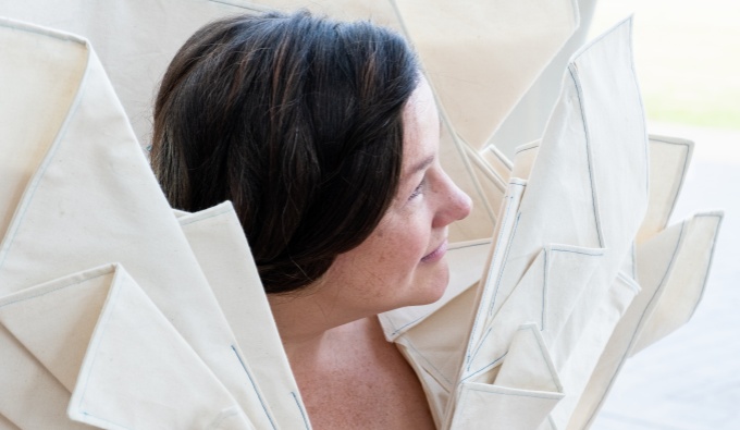 An head shot of Lisette from above. She has brown hair tied back, her head is turned to the side and large pleats on the shoulders of her linen dress fan outwards.