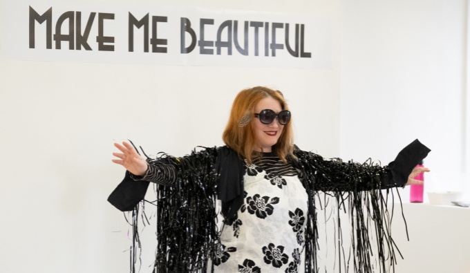 Stav stands in front of a white wall with black text; Make Me Beautiful. They wear sunglasses, a black fringed jacket and a clack and white floral top. 