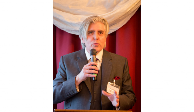 David Hevey is standing at a podium and holding a microphone, behind him are red and cream curtains. He is wearing a dark grey suit, with a red handkerchief in his breast pocket. 