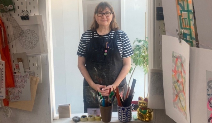 Emma wears a stripy blue and white t-shirt, dungarees and has shoulder length hair and wears dark rimmed glasses. They stand in a doorway, the light behind them. In front is a table with art equipment.