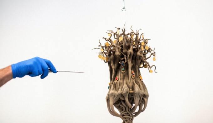 The top tier of a cake depicting a tangle of roots, suspended from the ceiling of a white gallery space. A hand wearing a blue latex glove holds a sharp metal object towards it, as if to cut or lance the cake,