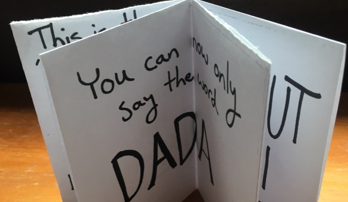 A white paper booklet with black text is open and stood upright. The word DADA is prominent.
