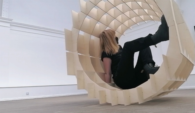  Situated within a large white gallery space, Sam wears all black and sits within the wooden cylindrical framework of one of her pieces.