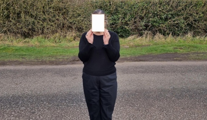 A person wearing drk clothing stands in front of a road with a green hedge beyond. They hold a white piece of paper in front of their face. 