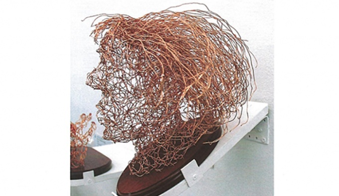 The profile view of a 3D copper wire sculpture of the artist's head, placed upon a dark wooden base.