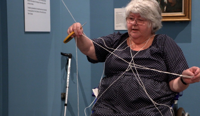 Jennette is seated in a wheelchair within a blue gallery space. She holds a long piece of string that also drapes across her shoulders and torso as she works.