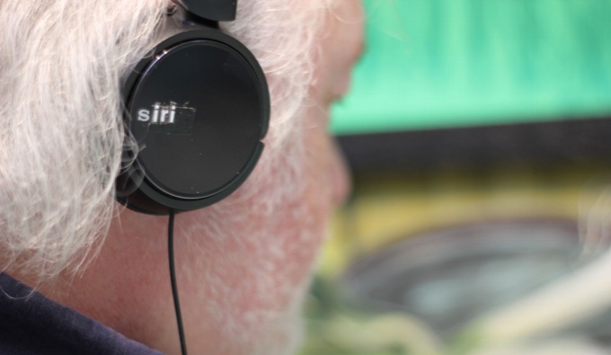 A close up of the artist wearing black headphones. He has white hair and white stubble.