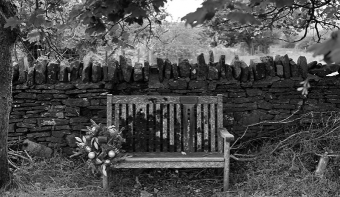 A black and white photograph of a wooden bench against a traditional dry stone wall.  Above is a leafy canopy of plane trees. The grass around the feet of the  bench is long and untamed and attached to the arm of the bench is a large floral arrangement.