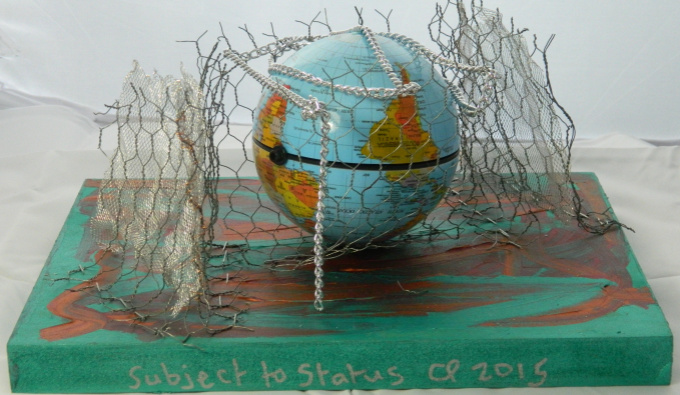 An upside down globe surrounded by chicken wire ontop of a green and orange painted canvas with the words 'Subject to Status CP 2015' painted on the side of it.