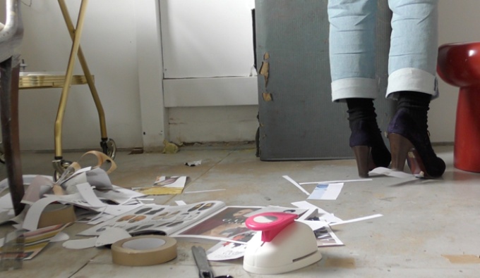 A room with flaking paint, a wooden ladder and cut up, discarded paper across the floor. The artist's feet are just in shot and wear smart dark chunky healed shoes and jeans with rolled up lower leg. 