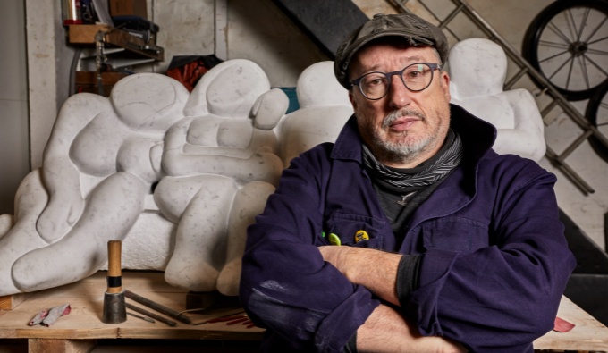 Tony Heaton wears glasses, a cap and a blue jacket. He stands in front of a large sculpture. partially obscured from view as he looks at the camera.