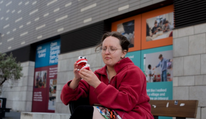 Alice wears a red jacket, dark jeans and a white t-shirt. She sits outside and holds a toy set of false teeth with eyes upon the top. 