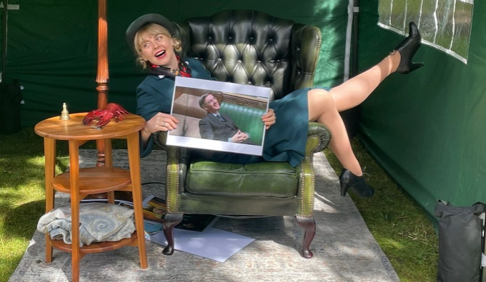 Dora wears a vintage style green skirt suit with a straw hat. She is smiling as she lies across a green leather armchair. In her hand she holds an image of a politician in the house of commons stood in front of the green leather benches. 