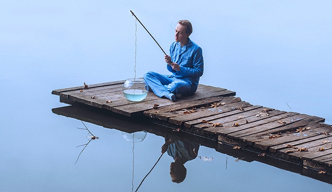 A figure sat at the end of a wooden jetty, surrounded by calm water. They wear a blue jacket and trousers and have cropped blonde hair. They are holding a wooden fishing rod, the line of which dangles into a round glass fish bowl.