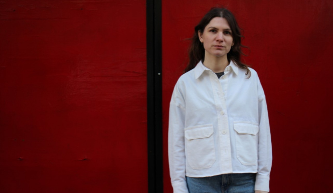 Hannah Wallis stands to the right hand side in front of a plain dark red background. She wears a white shirt and blue jeans and faces the camera.